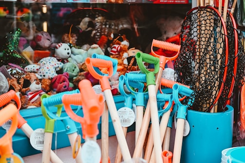 A vibrant collection of beach toys including shovels with colorful handles and nets in a bucket, displayed alongside a variety of plush toys inside a shop window.
