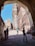 An architectural passageway frames a view of a historical fort made of sandstone with intricate carvings. The clear blue sky enhances the grandeur of the fort, while a group of people, including tourists and locals, can be seen walking or resting in the shaded area.