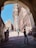 An architectural passageway frames a view of a historical fort made of sandstone with intricate carvings. The clear blue sky enhances the grandeur of the fort, while a group of people, including tourists and locals, can be seen walking or resting in the shaded area.