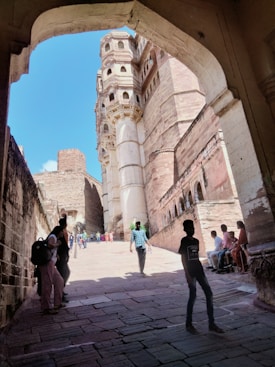 An architectural passageway frames a view of a historical fort made of sandstone with intricate carvings. The clear blue sky enhances the grandeur of the fort, while a group of people, including tourists and locals, can be seen walking or resting in the shaded area.