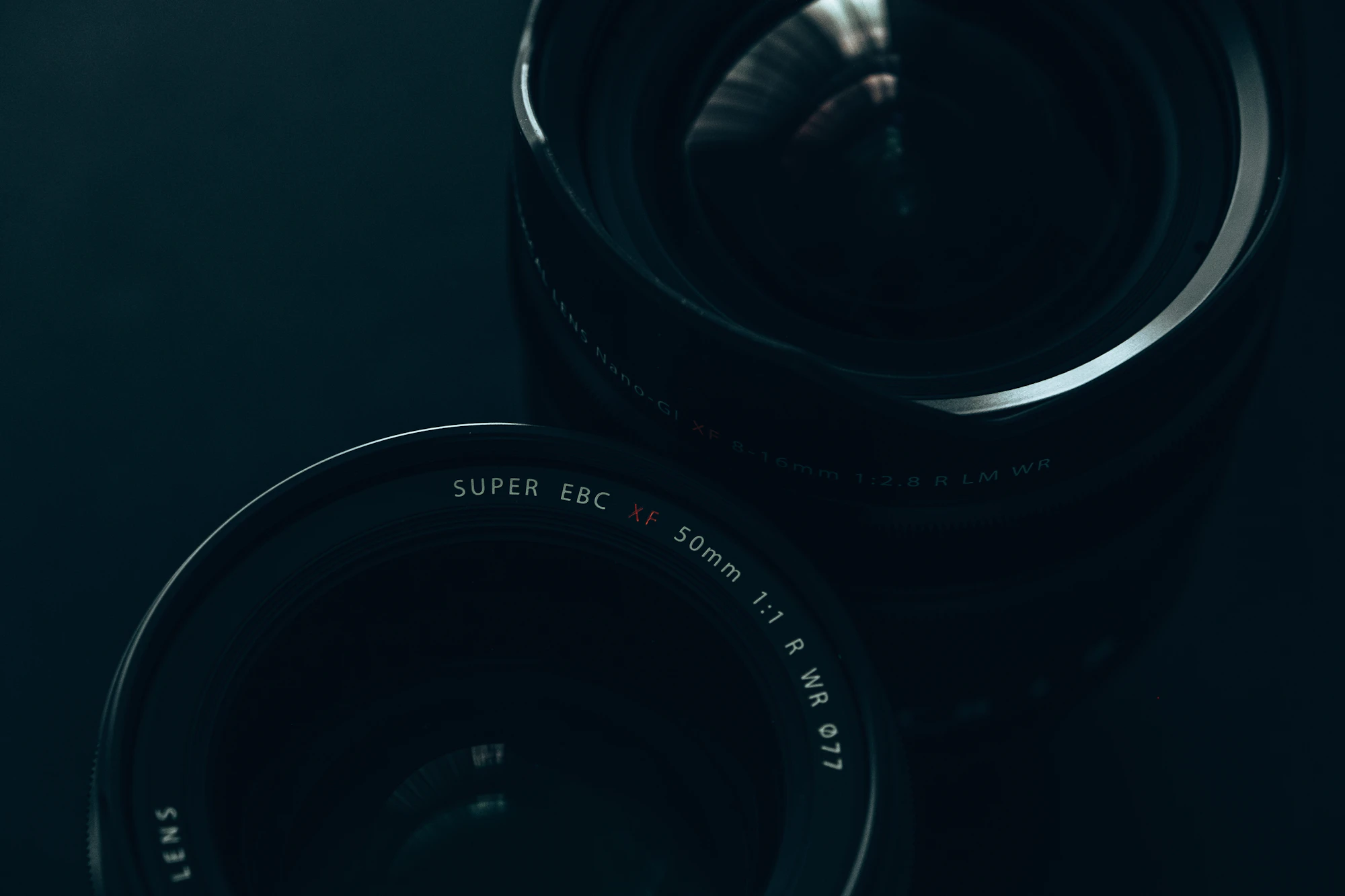 A cinema prime lens against deep black, a single edge of light along its barrel — the instrument, not the operator.