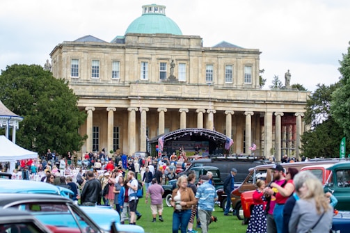 A large gathering of people is in front of a grand, classical-style building with a green dome. An outdoor event seems to be taking place with American flags displayed and various vintage cars parked around the area. The crowd includes a diverse group of people, some of whom are dressed in retro or vintage clothing.
