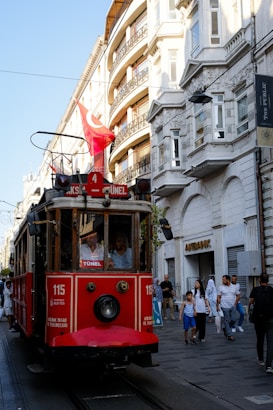 A vintage red tram is moving along a street lined with European-style buildings. Several pedestrians are walking nearby, and a Turkish flag is mounted on top of the tram. The ambient lighting suggests it is late afternoon with shadows falling across the scene.