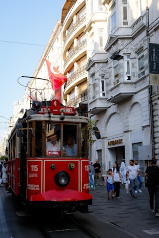 A vintage red tram is moving along a street lined with European-style buildings. Several pedestrians are walking nearby, and a Turkish flag is mounted on top of the tram. The ambient lighting suggests it is late afternoon with shadows falling across the scene.