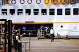 A large ferry docked at a port with multiple people standing near the entrance. The ferry is white with a yellow stripe and circular windows. Life buoys are mounted along the upper deck railing. One person is seen handling a heavy rope, possibly securing the vessel.