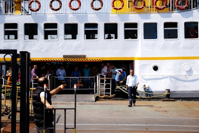 A large ferry docked at a port with multiple people standing near the entrance. The ferry is white with a yellow stripe and circular windows. Life buoys are mounted along the upper deck railing. One person is seen handling a heavy rope, possibly securing the vessel.