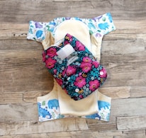 A reusable cloth diaper with a vibrant floral pattern on an outer layer is laid out. Beneath it is another diaper with a design featuring various colorful animal illustrations such as elephants and dinosaurs. These are set against a rustic wooden background.