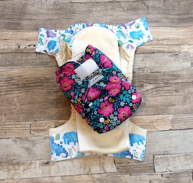 A reusable cloth diaper with a vibrant floral pattern on an outer layer is laid out. Beneath it is another diaper with a design featuring various colorful animal illustrations such as elephants and dinosaurs. These are set against a rustic wooden background.