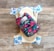 A reusable cloth diaper with a vibrant floral pattern on an outer layer is laid out. Beneath it is another diaper with a design featuring various colorful animal illustrations such as elephants and dinosaurs. These are set against a rustic wooden background.