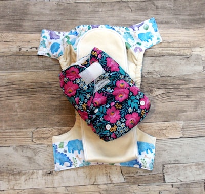 A reusable cloth diaper with a vibrant floral pattern on an outer layer is laid out. Beneath it is another diaper with a design featuring various colorful animal illustrations such as elephants and dinosaurs. These are set against a rustic wooden background.