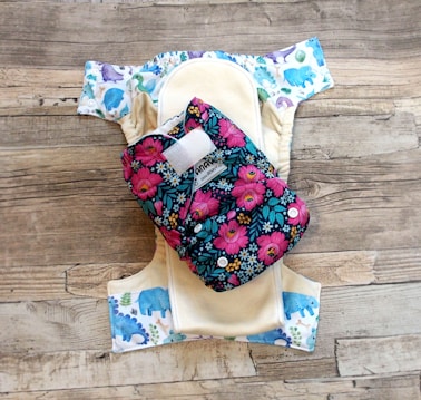 A reusable cloth diaper with a vibrant floral pattern on an outer layer is laid out. Beneath it is another diaper with a design featuring various colorful animal illustrations such as elephants and dinosaurs. These are set against a rustic wooden background.