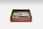 An ornately decorated box with an intricate red design holds a rectangular stone ink slab and a small, round lid with gold accents. The items are neatly placed inside the box, which features a black interior.