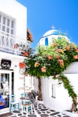 A charming street scene featuring a white-washed building with a blue dome typical of Cycladic architecture. Vibrant orange flowers cascade down a trellis, adding a splash of color. The cobblestone path leads past a small café area with turquoise chairs and tables, creating an inviting and picturesque setting.