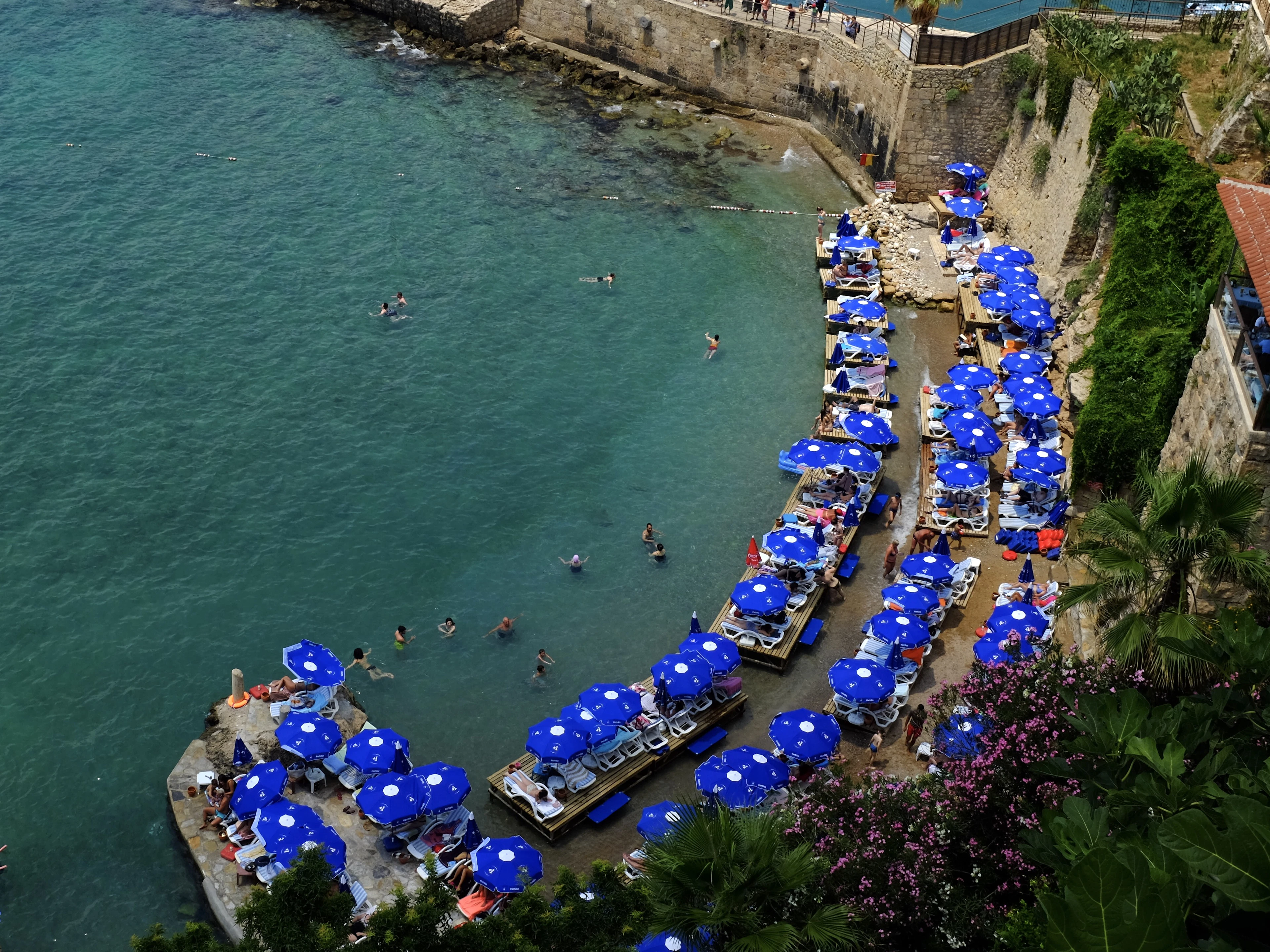 Blue beach umbrellas line a pebble cove built into Antalya's old city walls, swimmers in turquoise water below.