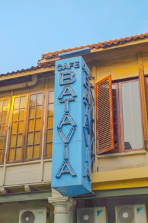 A vintage building with large windows and wooden shutters is adorned with a vertical blue sign that reads 'Cafe Batavia' in bold, large letters. The building has an old-world charm with warm golden light reflecting off the window panes, suggesting either sunrise or sunset.
