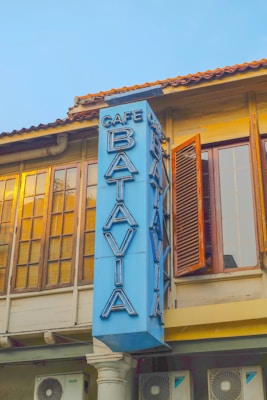 A vintage building with large windows and wooden shutters is adorned with a vertical blue sign that reads 'Cafe Batavia' in bold, large letters. The building has an old-world charm with warm golden light reflecting off the window panes, suggesting either sunrise or sunset.