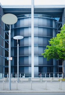 A modern, cylindrical building with dark horizontal blinds, framed by two vertical structures. The facade includes a tree with lush green leaves and a few street lamps in the foreground. A bicycle is parked at the base.