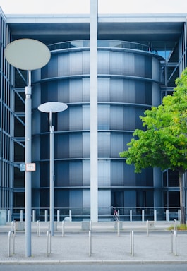 A modern, cylindrical building with dark horizontal blinds, framed by two vertical structures. The facade includes a tree with lush green leaves and a few street lamps in the foreground. A bicycle is parked at the base.
