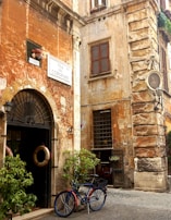 A rustic street corner features an old, weathered building with orange and beige walls and shuttered windows. A vintage sign reads 'Via dei Coronari'. A blue bicycle with an orange accent is parked against the building, and two life preservers hang on the door. Potted plants add greenery to the scene, and a small statue in an alcove is visible above a window.