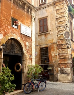 A rustic street corner features an old, weathered building with orange and beige walls and shuttered windows. A vintage sign reads 'Via dei Coronari'. A blue bicycle with an orange accent is parked against the building, and two life preservers hang on the door. Potted plants add greenery to the scene, and a small statue in an alcove is visible above a window.