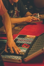 A person reaches into a poker chip case on a red felt table, while another person holds playing cards. The scene is dimly lit, creating a warm and engaged atmosphere.