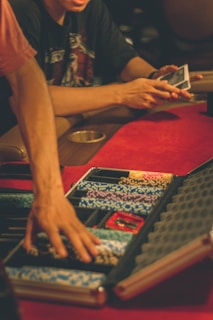 A person reaches into a poker chip case on a red felt table, while another person holds playing cards. The scene is dimly lit, creating a warm and engaged atmosphere.