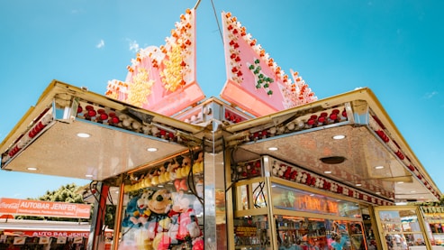 A colorful amusement park stall featuring bright red, white, green, and yellow lights. The stall displays a variety of plush toys, including various animals, which are visible through the glass-front panels. The structure has a geometric design with metallic accents and is set against a clear blue sky.