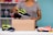 A person is placing a sneaker into a cardboard box. The sneaker is navy blue with bright green accents. The person is wearing a gray shirt and is standing at a table with another sneaker and folded clothes on it. In the background, shelves hold various items including dumbbells and colorful storage boxes against a teal wall.