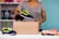 A person is placing a sneaker into a cardboard box. The sneaker is navy blue with bright green accents. The person is wearing a gray shirt and is standing at a table with another sneaker and folded clothes on it. In the background, shelves hold various items including dumbbells and colorful storage boxes against a teal wall.