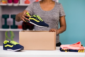 A person is placing a sneaker into a cardboard box. The sneaker is navy blue with bright green accents. The person is wearing a gray shirt and is standing at a table with another sneaker and folded clothes on it. In the background, shelves hold various items including dumbbells and colorful storage boxes against a teal wall.