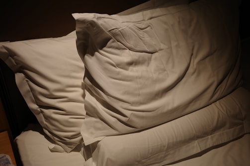 Two white pillows resting on a bed with soft lighting casting shadows. The bedding appears smooth and neatly arranged, creating a cozy and inviting atmosphere.