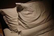 Two white pillows resting on a bed with soft lighting casting shadows. The bedding appears smooth and neatly arranged, creating a cozy and inviting atmosphere.