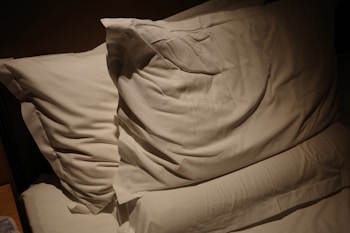 Two white pillows resting on a bed with soft lighting casting shadows. The bedding appears smooth and neatly arranged, creating a cozy and inviting atmosphere.