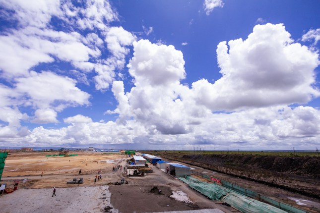 A wide expanse of land with a construction site, featuring a partially built structure, machinery, and people scattered across the area. The sky is filled with large, fluffy clouds against a vibrant blue backdrop.