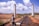 Workers are engaged in construction activities on a railway track, with two prominent pillars in the background. Several individuals can be seen working along the track, including one person climbing a pole. The area is set in a vast landscape with hills in the distance, and the sky is dotted with fluffy clouds.