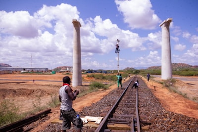 Workers are engaged in construction activities on a railway track, with two prominent pillars in the background. Several individuals can be seen working along the track, including one person climbing a pole. The area is set in a vast landscape with hills in the distance, and the sky is dotted with fluffy clouds.