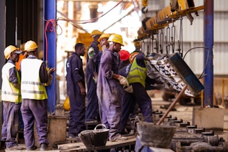 A group of workers wearing yellow safety helmets and reflective vests engage in industrial activities inside a factory setting. Some are observing while others are actively operating machinery as a large piece of equipment is lifted by mechanical hoists.