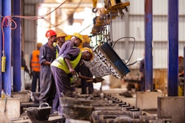Several workers are actively engaged in an industrial setting, wearing protective gear such as helmets and vests. They are operating machinery or handling materials suspended from an overhead crane. The environment appears to be a factory or workshop with metal beams and industrial equipment.