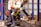 Several workers are actively engaged in an industrial setting, wearing protective gear such as helmets and vests. They are operating machinery or handling materials suspended from an overhead crane. The environment appears to be a factory or workshop with metal beams and industrial equipment.