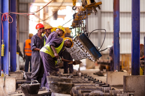 Several workers are actively engaged in an industrial setting, wearing protective gear such as helmets and vests. They are operating machinery or handling materials suspended from an overhead crane. The environment appears to be a factory or workshop with metal beams and industrial equipment.