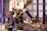 Several workers are actively engaged in an industrial setting, wearing protective gear such as helmets and vests. They are operating machinery or handling materials suspended from an overhead crane. The environment appears to be a factory or workshop with metal beams and industrial equipment.