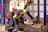 Several workers are actively engaged in an industrial setting, wearing protective gear such as helmets and vests. They are operating machinery or handling materials suspended from an overhead crane. The environment appears to be a factory or workshop with metal beams and industrial equipment.