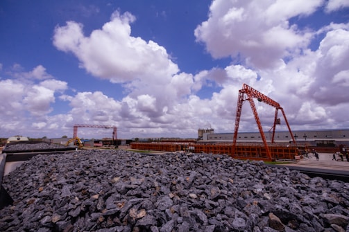 A construction site featuring large mounds of rocks in the foreground and red metal structures in the background. The sky is filled with numerous fluffy white clouds against a bright blue backdrop. Buildings and construction vehicles are visible in the distance.