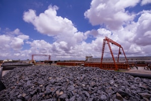 A construction site featuring large mounds of rocks in the foreground and red metal structures in the background. The sky is filled with numerous fluffy white clouds against a bright blue backdrop. Buildings and construction vehicles are visible in the distance.