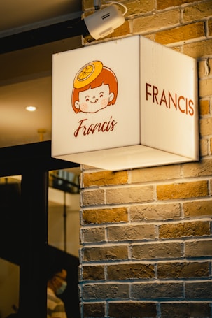 A lighted sign on a brick wall features a cartoon face with a citrus fruit on top of the head and the name 'Francis' written below. The sign is illuminated and mounted on the corner of a building. Above the sign, a security camera is visible.