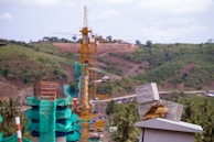 A construction site set amid a hilly, green landscape features several cranes and partially constructed structures clad in green netting. In the foreground, there are concrete slabs with handwritten notes and a pair of work gloves resting on top of a flat surface. The background shows rolling hills and scattered trees under a partly cloudy sky.