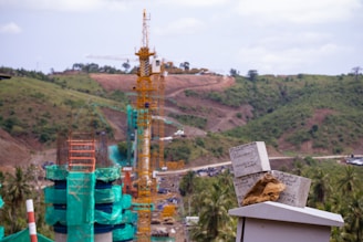 A construction site set amid a hilly, green landscape features several cranes and partially constructed structures clad in green netting. In the foreground, there are concrete slabs with handwritten notes and a pair of work gloves resting on top of a flat surface. The background shows rolling hills and scattered trees under a partly cloudy sky.