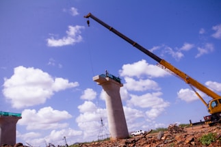 A construction site features large concrete pillars with a crane lifting an object. The sky is bright blue with scattered white clouds, and there is a truck and debris in the background.