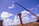 A construction site features large concrete pillars with a crane lifting an object. The sky is bright blue with scattered white clouds, and there is a truck and debris in the background.