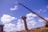 A construction site features large concrete pillars with a crane lifting an object. The sky is bright blue with scattered white clouds, and there is a truck and debris in the background.